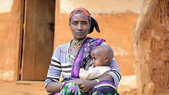 Eine Frau sitzt vor einer Lehmhütte, sie hält ein Kleinkind im Arm und schaut ernst in die Kamera