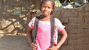 Mädchen mit Schulrucksack steht vor einer Lehmziegelmauer in einem afrikanischen Dorf und lächelt in die Kamera