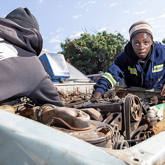 Zwei junge Frauen in Overall beugen sich über den Motorblock eines alten Autos