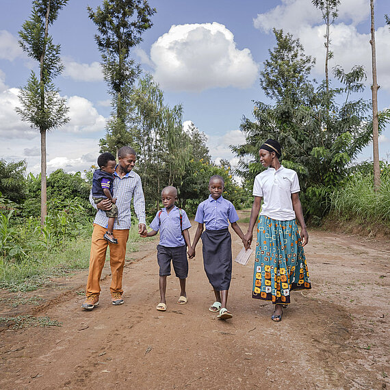 Familie geht gemeinsam auf einem Feldweg in Ruanda Eine Familie aus Ruanda geht gemeinsam auf einem Feldweg: Ein Mann trägt ein Kleinkind auf dem Arm, während zwei Kinder und eine Frau die Hand halten. Im Hintergrund sind Bäume und ein grüner Landschaftsteil zu sehen.