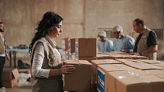 Eine Frau steht in einem Lagerhaus. Sie verschließt eine Box, in auf der das Plan-International-Logo ist und stellt sie zu vielen weiteren Boxen.