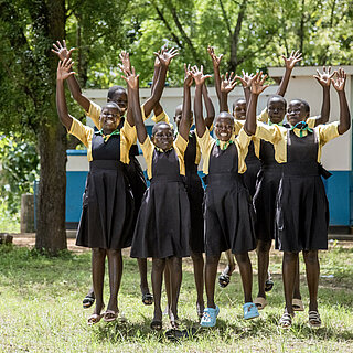 Neues aus der Stiftung Gruppe von fröhlichen Schülerinnen in Schuluniformen springt mit erhobenen Armen auf einer Wiese vor einem Schulgebäude in Uganda.