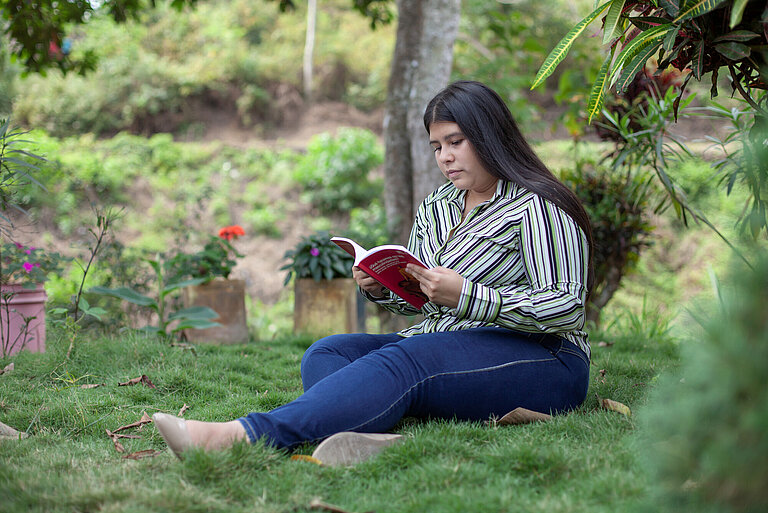 Eine junge Frau mit langen, dunklen Haaren sitzt auf dem Rasen und liest ein Buch