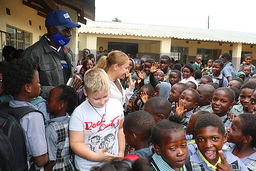 Eine große Gruppe sambischer Schulkinder begrüßt eine Familie und einen Projektmitarbeiter auf dem Schulhof in Sambia; alle sind fröhlich und interessiert, Begegnung und Austausch stehen im Mittelpunkt.