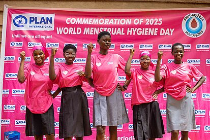 Fünf junge Frauen stehen nebeneinander und halten ihre rechte Faust nach oben. Sie tragen ein pinkes T-Shirt mit Plan-Logo und einem Herzen. Hinter ihnen ist eine Plakatwand auf der "World Menstrual Hygiene Day" steht.