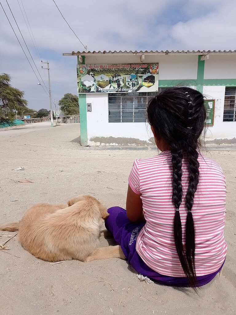 Ein junges Mädchen sitzt mit dem Rücken zur Kamera auf der Straße, neben ihm liegt ein kleiner Hund