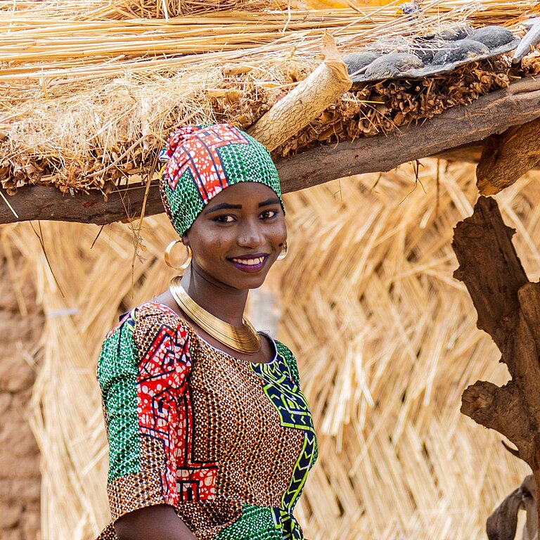 Eine junge Frau aus Nigeria steht draußen, sie trägt ein buntes Kleid und ein Tuch um das Haar gewickelt und lächelt in die Kamera
