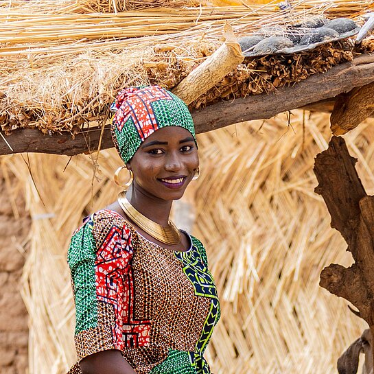 Eine junge Frau aus Nigeria steht draußen, sie trägt ein buntes Kleid und ein Tuch um das Haar gewickelt und lächelt in die Kamera