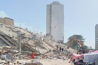 Menschen und Hilfskräfte bewegen sich zwischen den Trümmern eines eingestürzten Gebäudes in La Guaira, Venezuela; im Hintergrund arbeiten Bagger und weitere Gebäude sind sichtbar.