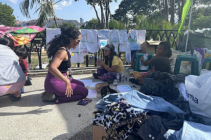 Eine Helferin der Partnerorganisation Tinta Violeta betreut mehrere Kinder, die im Freien sitzen und malen; im Hintergrund hängen Kinderzeichnungen an einem Zaun.