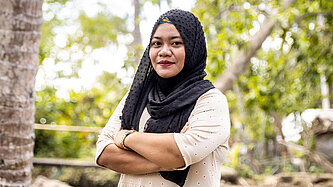 Farhana steht mit verschränkten Armen und lächelt selbstbewusst in die Kamera. Sie trägt ein cremefarbenes Kleid und ein schwarzes Kopftuch.