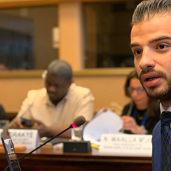 Karim sitzt in einer Konferenz und spricht in ein Mikrofon.