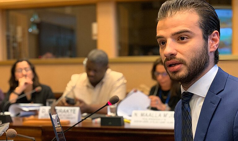 Karim sitzt in einer Konferenz und spricht in ein Mikrofon.