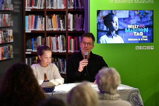 Schauspieler Tim Bergmann, mit einem Mikrofon in der Hand, und eine Schülerin sitzen für eine Lesung an einem Tisch