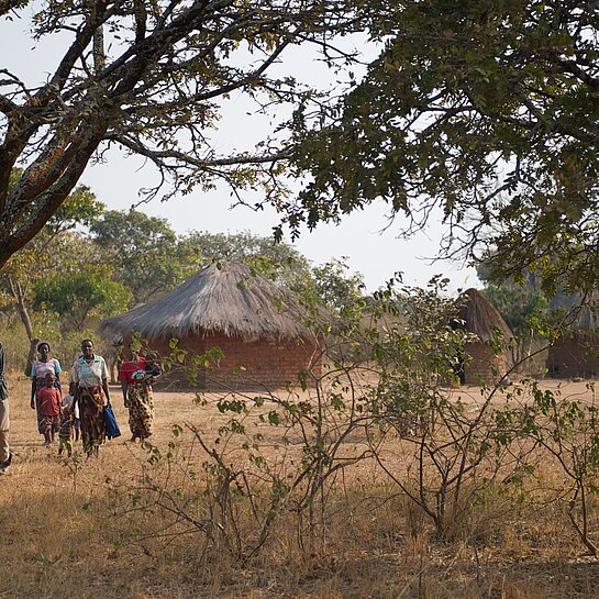 Mehrere Menschen gehen durch eine ländliche, trockene Landschaft in Sambia, vorbei an Bäumen und traditionellen Rundhütten mit Strohdach.