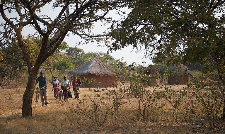 Mehrere Menschen gehen durch eine ländliche, trockene Landschaft in Sambia, vorbei an Bäumen und traditionellen Rundhütten mit Strohdach.