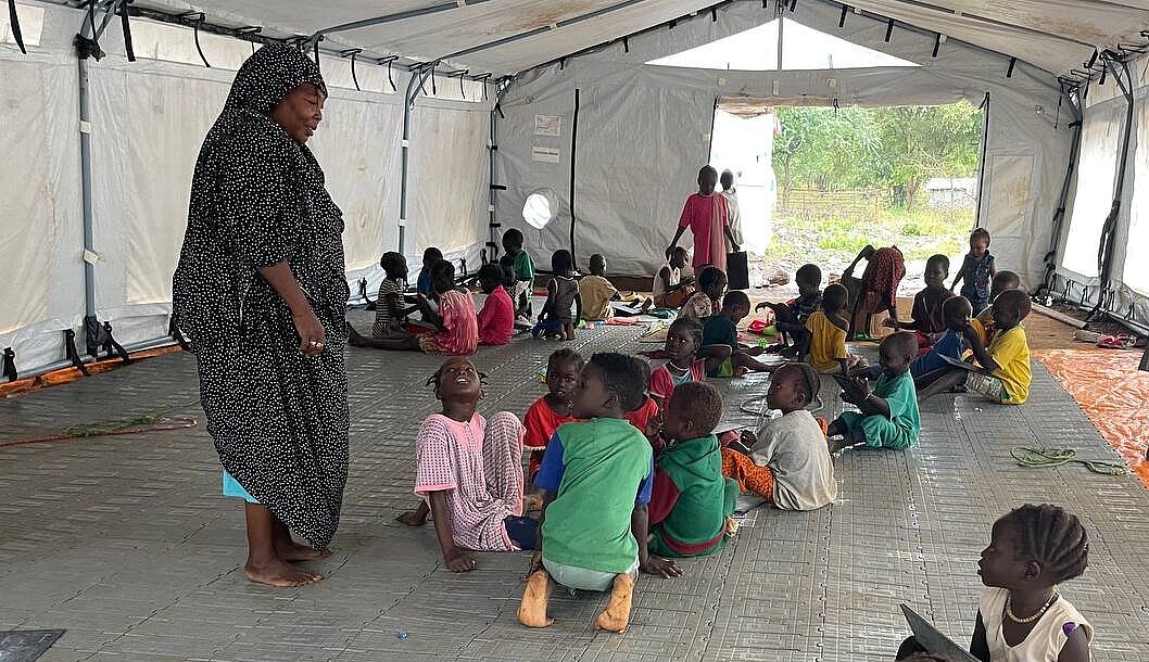 Viele Kinder sitzen in einem großen Zelt auf dem Boden, eine Frau steht in der Mitte und spricht mit einer Gruppe Kinder
