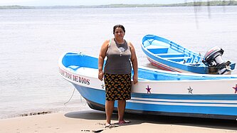 Eine Frau steht am Strand, im Hintergrund ist ein Fischerboot am Ufer zu sehen.