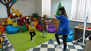 Geflüchtete Kinder tanzen in einem bunten Klassenzimmer im Rahmen einer Nothilfeaktion im Kontext des Ukraine-Kriegs. Das Klassenzimmer ist hell und freundlich gestaltet, mit weichen Sitzsäcken und einer fröhlichen Atmosphäre.