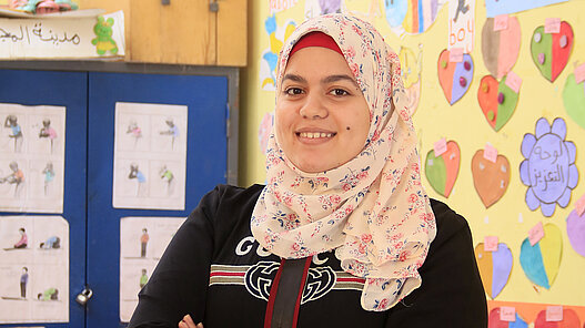 Eine junge Frau mit verschränkten Armen steht lächelnd in einem Klassenzimmer.