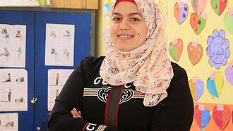 Eine junge Frau mit verschränkten Armen steht lächelnd in einem Klassenzimmer.