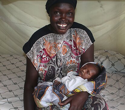 Eine Frau sitzt auf einem Bett mit gemusterten Bezügen und hält ein kleines Baby in ihren Armen. Sie trägt ein farbenfrohes Kleid mit detaillierten Mustern.