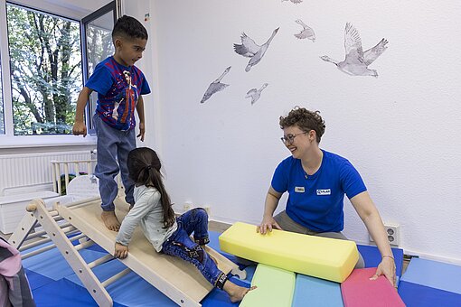 Zwei Kinder spielen auf einem Kletterdreieck in einem Raum, eine Frau in blauem T-Shirt spielt mit ihnen