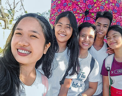 Fünf Jugendliche machen ein fröhliches Selfie im Freien bei sonnigem Wetter. Zwei Mädchen stehen vorne, dahinter drei weitere Jugendliche, ein bunter Sternen-Schirm bietet etwas Schatten. Alle lächeln in die Kamera, sie tragen Freizeitkleidung und Rucksäcke.