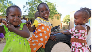  Vier kleine Mädchen spielen lachend draußen vor einem Reifen – Szene aus einem Dorf in Westafrika.