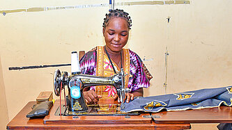 Eine junge Frau sitzt zufrieden an einer Nähmaschine und vernäht einen bunten Stoff
