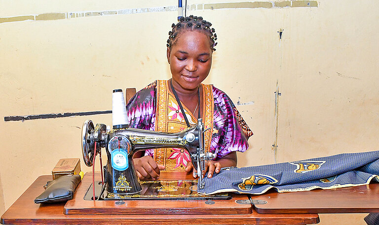 Eine junge Frau sitzt zufrieden an einer Nähmaschine und vernäht einen bunten Stoff