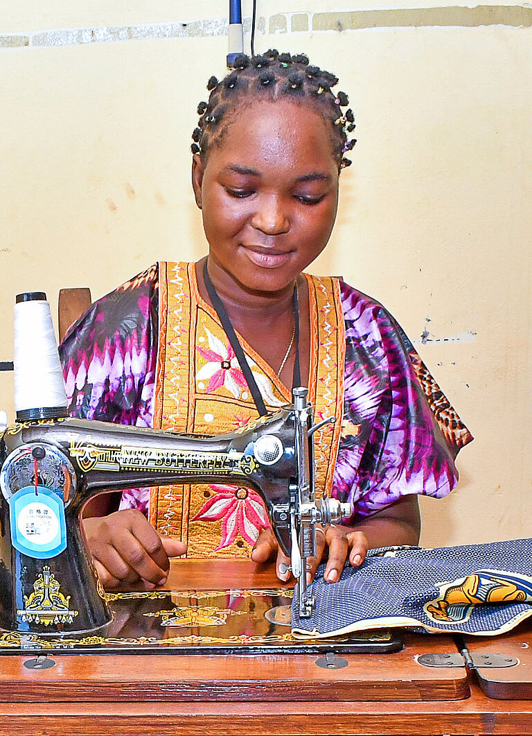 Eine junge Frau sitzt zufrieden an einer Nähmaschine und vernäht einen bunten Stoff