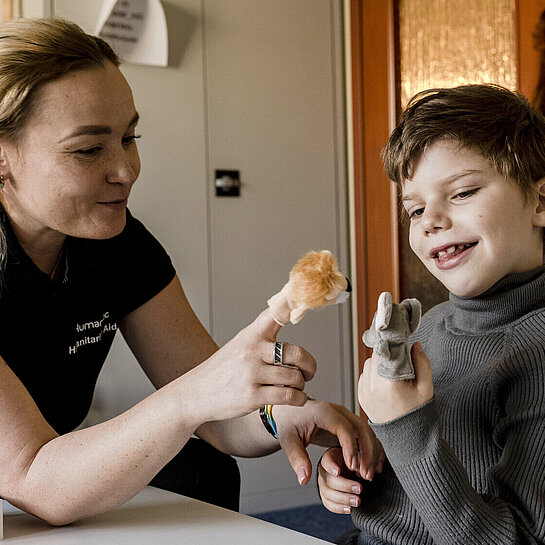 Eine Frau sitzt neben einem Jungen an einem Tisch. Die beiden spielen mit Fingerpuppen und lachen