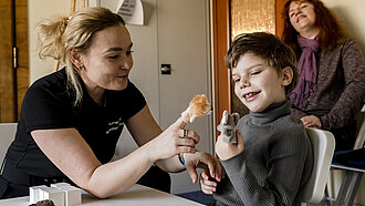 Eine Frau sitzt neben einem Jungen an einem Tisch. Die beiden spielen mit Fingerpuppen und lachen