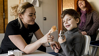 Eine Frau sitzt neben einem Jungen an einem Tisch. Die beiden spielen mit Fingerpuppen und lachen