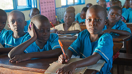 Mehrere Kinder sitzen in einer Klassensituation an Holzbänken. Sie tragen blaue Schuluniformen. Ein Kind im Vordergrund schreibt konzentriert in sein Heft, während ein anderes Kind sich über das Heft beugt. 