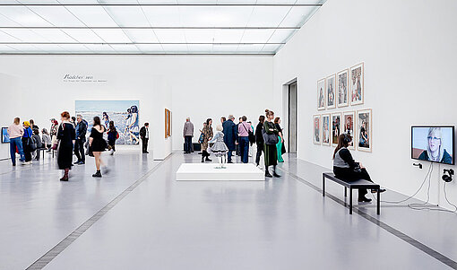 Personen laufen durch einen schneeweißen Raum, an dessen Wänden große Bilder und Fotos hängen