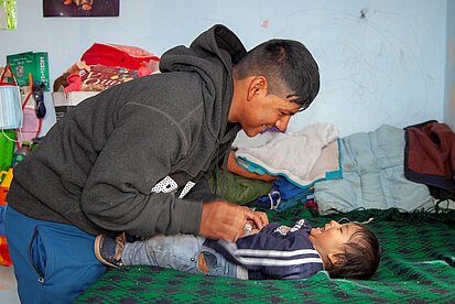Ein Erwachsener beugt sich über ein Kleinkind, das auf einem Bett mit grüner Decke liegt, und berührt spielerisch dessen Bauch; im Hintergrund liegen Kleidung und Gegenstände auf dem Bett.