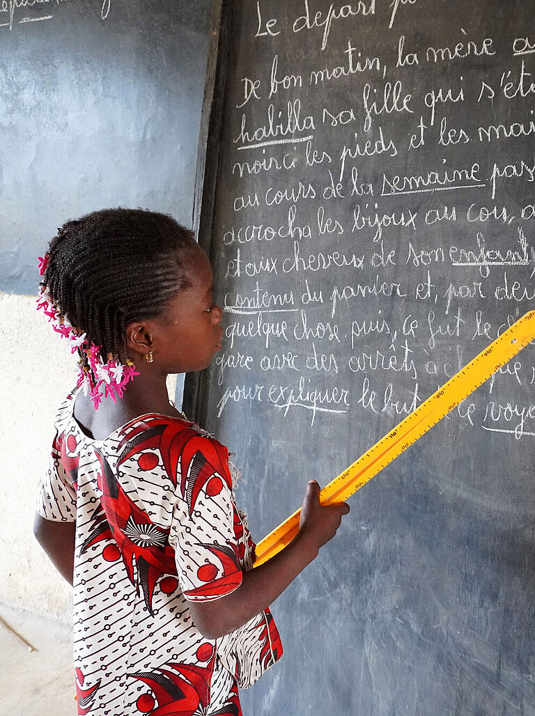 Ein Mädchen steht vor einer Tafel und zeigt mit dem Lineal auf Wörter, während sie sie vorliest.
