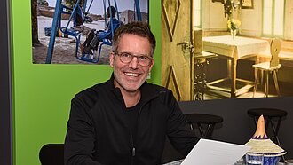 Schauspieler Tim Bergmann sitzt an einem Tisch und hält ein Blatt Papier in der Hand