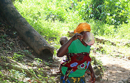 Eine liberische Frau trägt ein Kind auf dem Rücken, steht im Wald und trinkt aus einem Messbecher Wasser