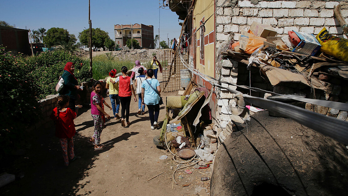 Mädchen laufen durch eine Gasse an einem Gebäude vorbei. Daneben liegt Sperrmüll und Abfall.
