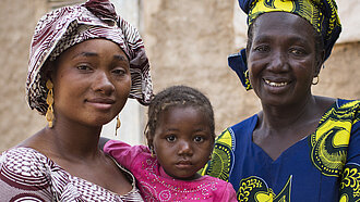 Drei Generationen afrikanischer Frauen, Mutter, Tochter und Großmutter, lächeln vor einem traditionellen Lehmhaus