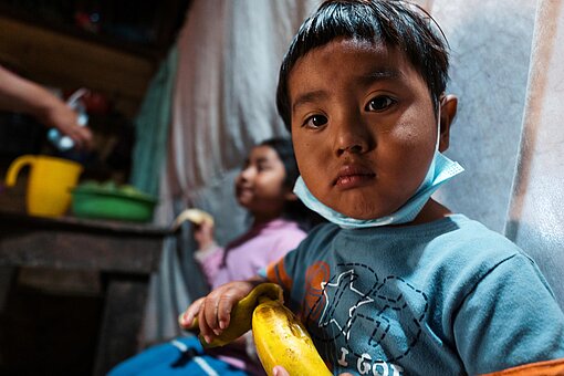 Ein kleiner Junge hält eine Banane in der Hand und schaut ernst in die Kamera.