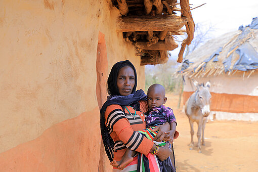 Eine Mutter steht mit ihrer einjährigen Tochter auf dem Arm vor einer Hütte