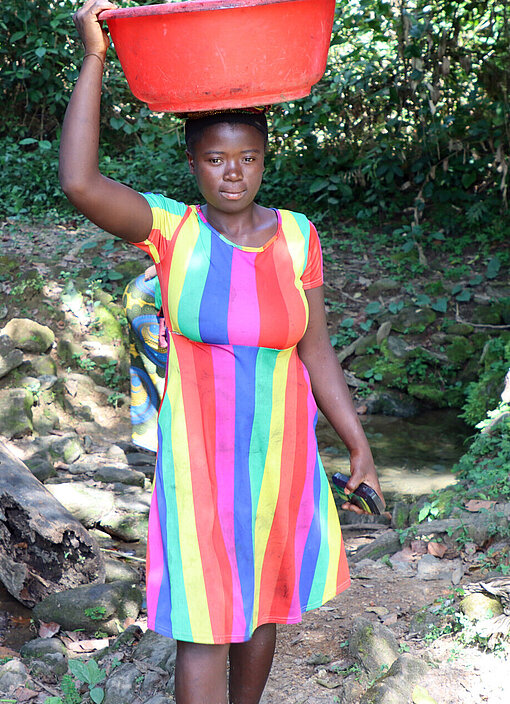 Ein liberianisches Mädchen in einem bunten Kleid trägt einen Wasserkanister auf dem Kopf
