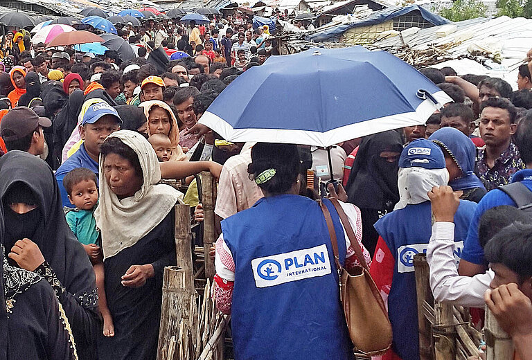 Plan-Teams stehen wartenden Rohingya-Familien gegenüber.