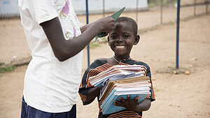 Lachendes ugandisches Kind trägt einen großen Stapel Schulhefte auf dem Arm, im Hintergrund eine weitere Person