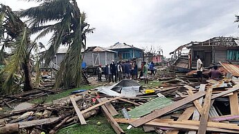Man sieht eine menge Holzplanken von abgedeckten Dächern auf dem Boden herumliegen, im Hintergrund umgewehte Palmen und zerstörte Häuserser