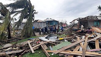 Man sieht eine menge Holzplanken von abgedeckten Dächern auf dem Boden herumliegen, im Hintergrund umgewehte Palmen und zerstörte Häuserser